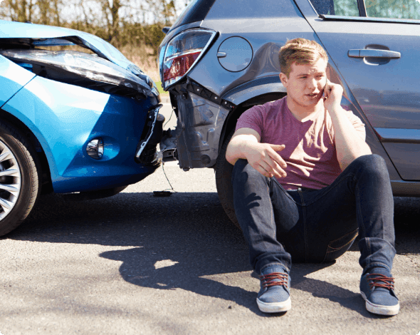 A person sitting on the road and speaking on the phone next to a car accident.