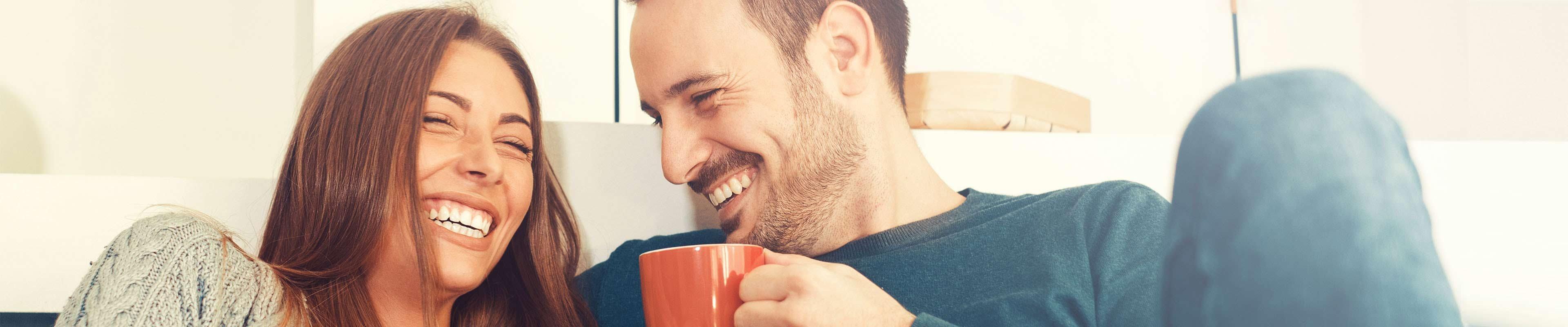 A brown-haired white woman sitting on the couch next to a brown-haired white man with short stubble laughing at a shared joke while the man holds a red coffee cup.