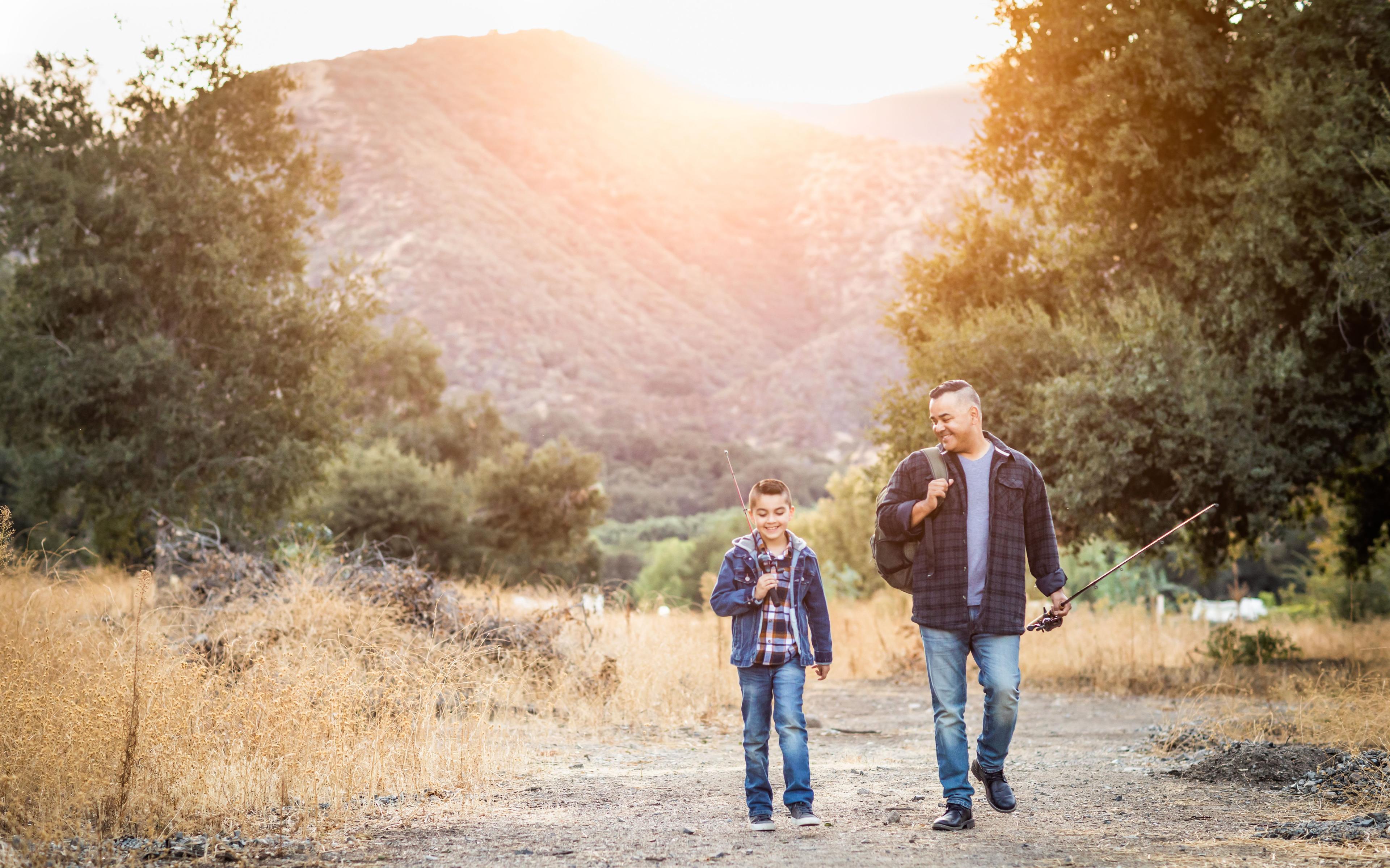 a man and a boy walking on a dirt road with trees and hills in the background